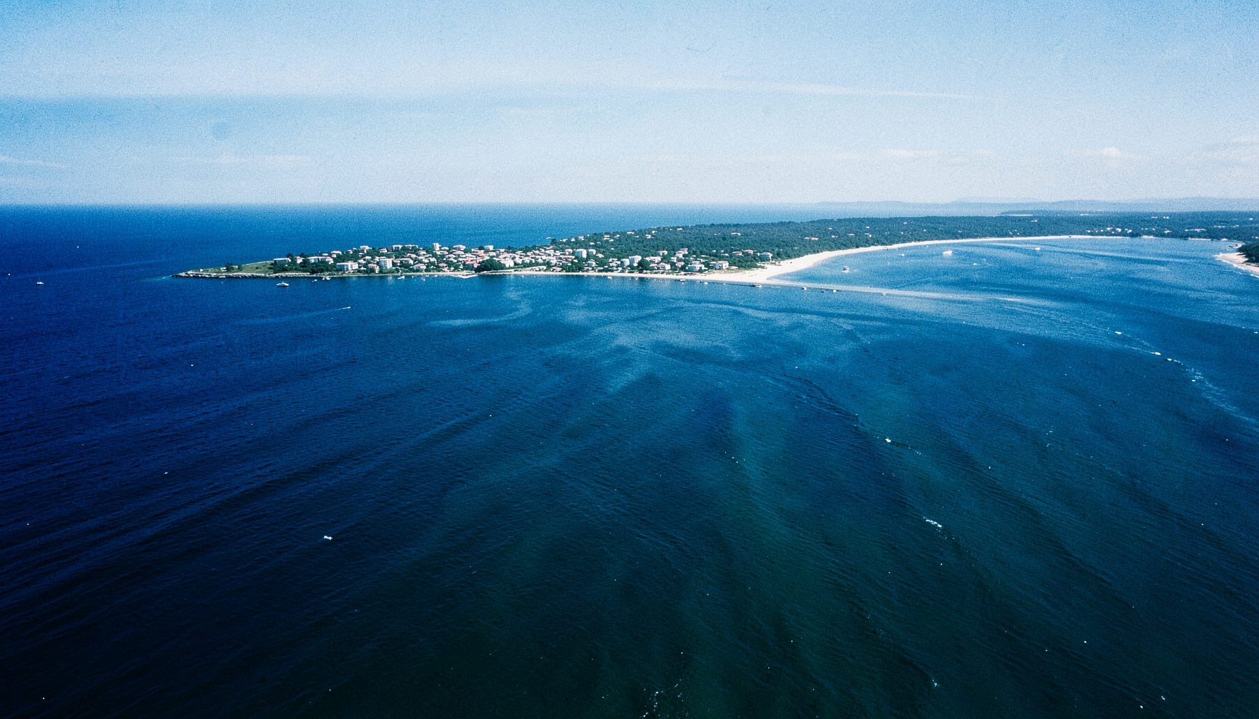 Aerial view of the Rockaway peninsula on a bright day, deep blue ocean on one side and calm bay waters on the other, white sand beaches stretching along the coast under a clear sky