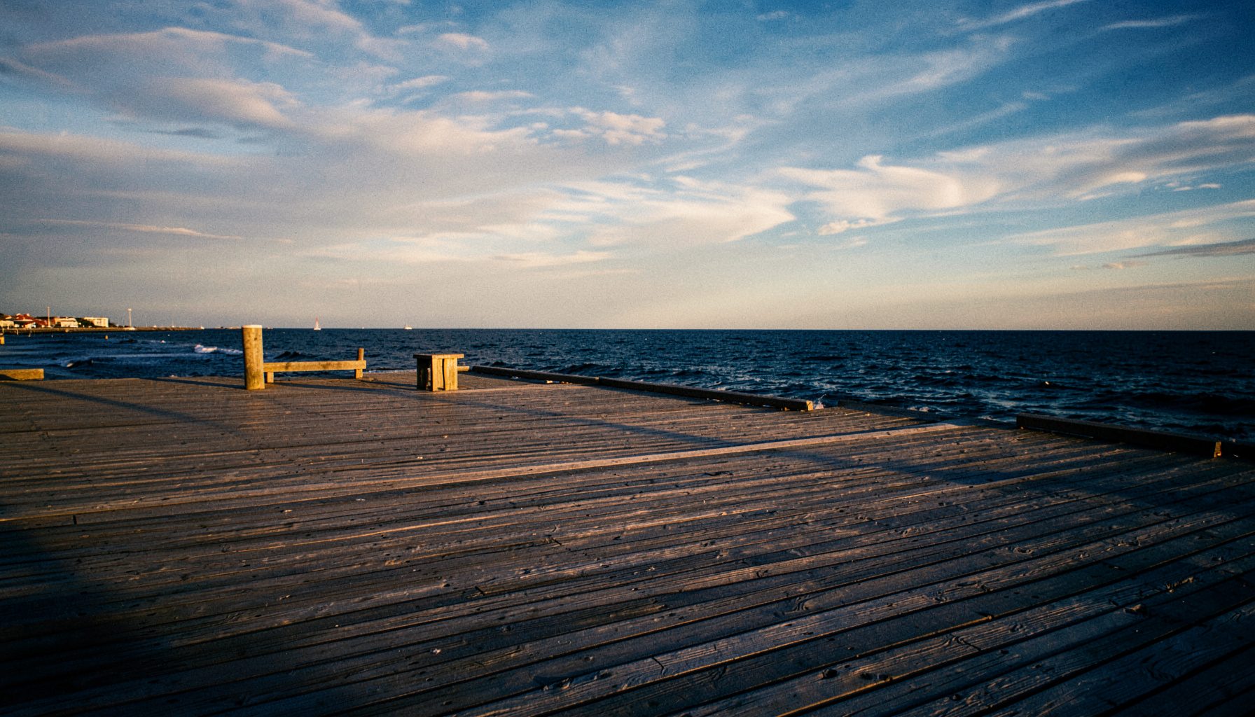 Wide photograph of a Rockaway boardwalk at golden hour, warm amber light on weathered wooden planks, deep navy ocean in the distance, soft clouds streaking the sky