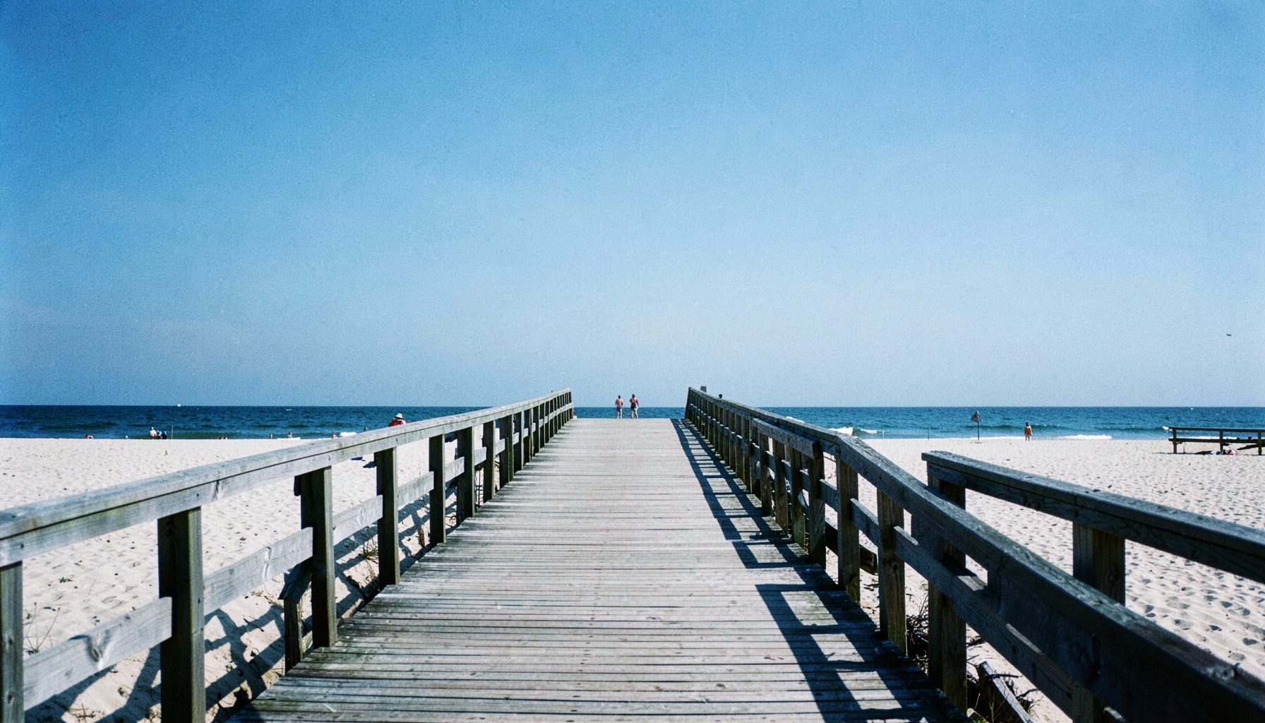 Wide view of a Rockaway beach boardwalk on a clear day, pale sand and soft blue sky meeting the ocean horizon