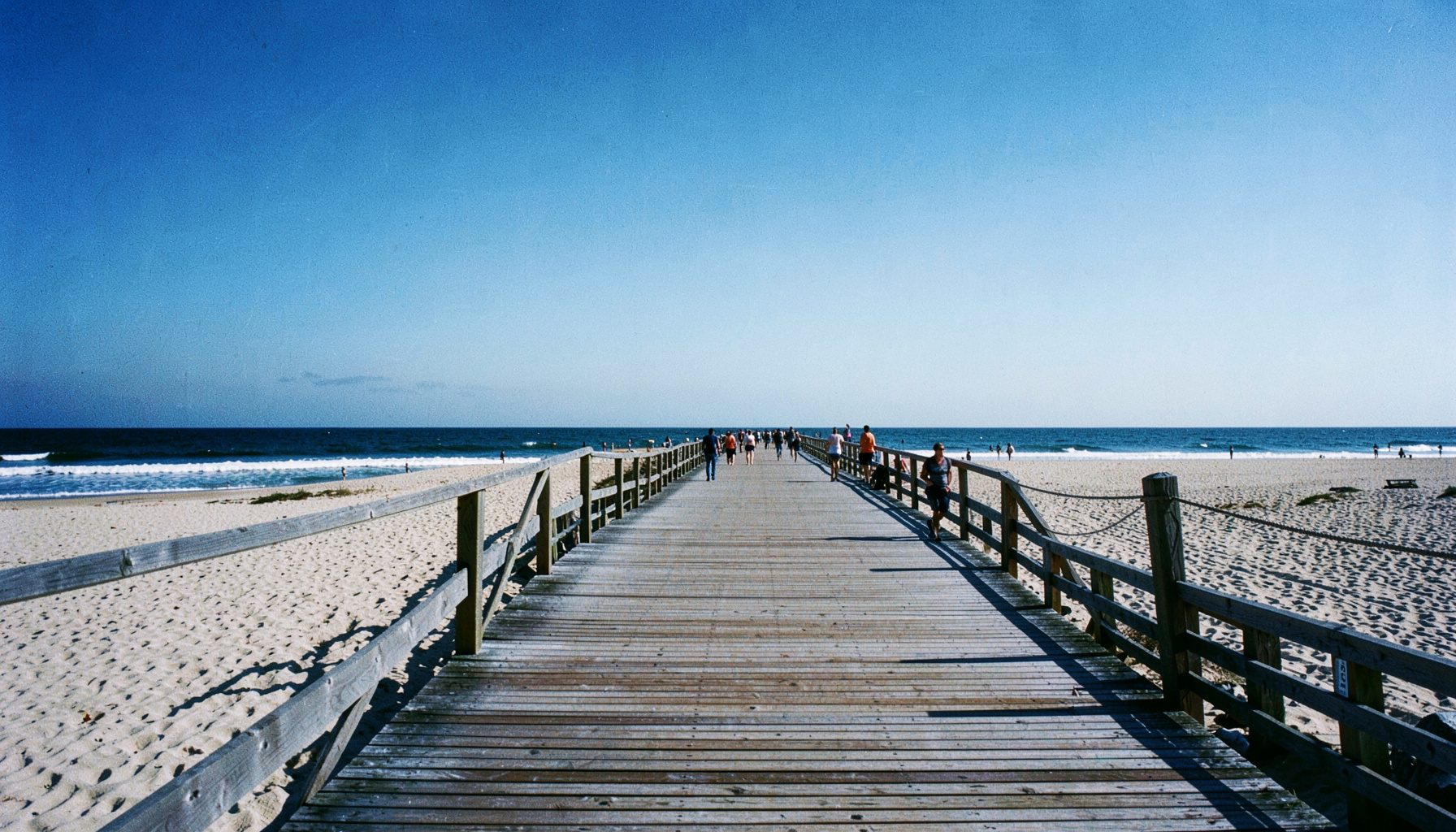 Wide view of the Rockaway Beach boardwalk on a clear day, with pale sand, blue sky, and gentle ocean waves in the distance